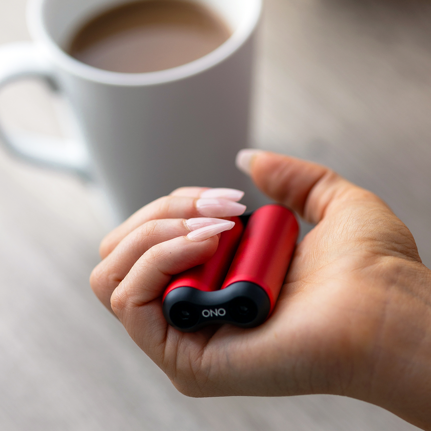 A hand with long, manicured nails holds a small, red ONO Roller Aluminum device near a white coffee mug on a light surface—perfect for fidgeting moments.