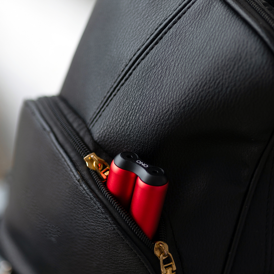 A pair of red wireless earbuds in a black charging case sits in the open front pocket of a black leather backpack with gold zippers—next to an ONO Roller Aluminum, ideal for discreet fidgeting on the move.