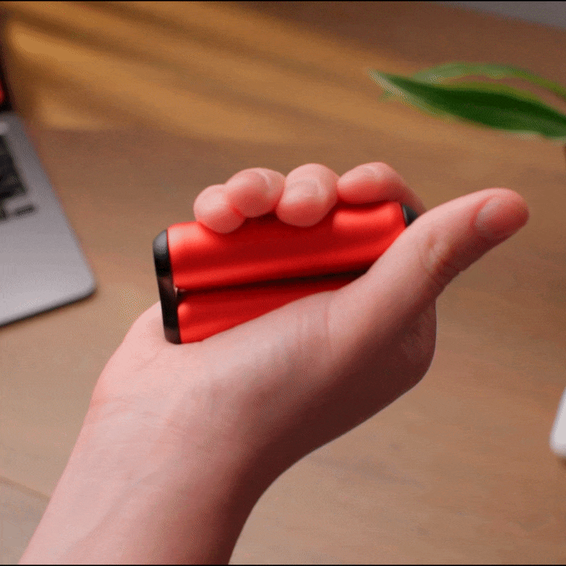 A hand squeezes a red grip strengthener on a wooden desk, with part of a laptop and green plant in the background—ideal for fidgeting or pairing with the ONO Roller Aluminum for stress relief.