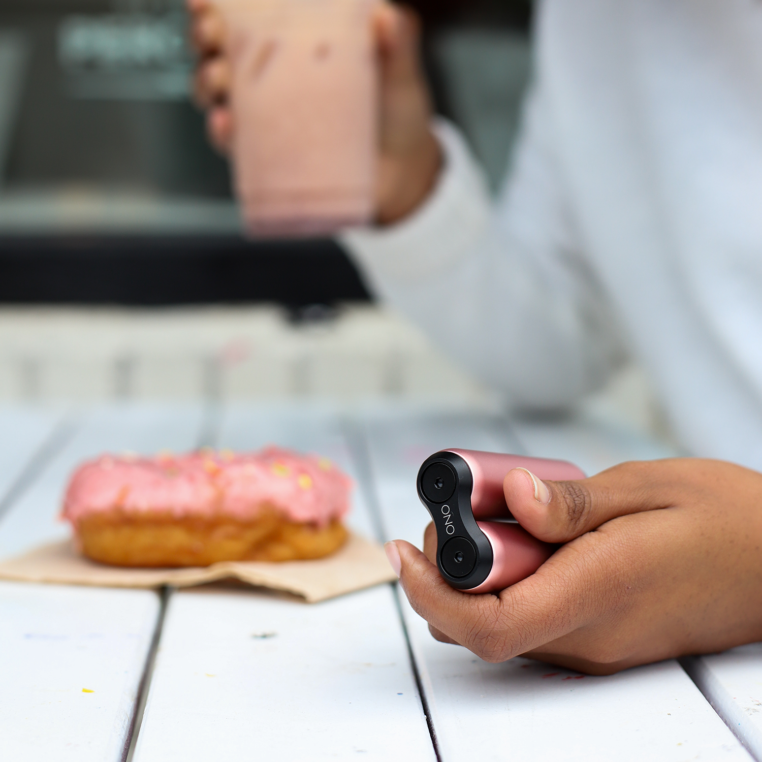 A person holds a pink ONO Roller Aluminum in one hand and a pink drink in the other, next to a pink frosted donut on a white wooden table. The sleek aluminum design makes it perfect for fidgeting while enjoying your treat.