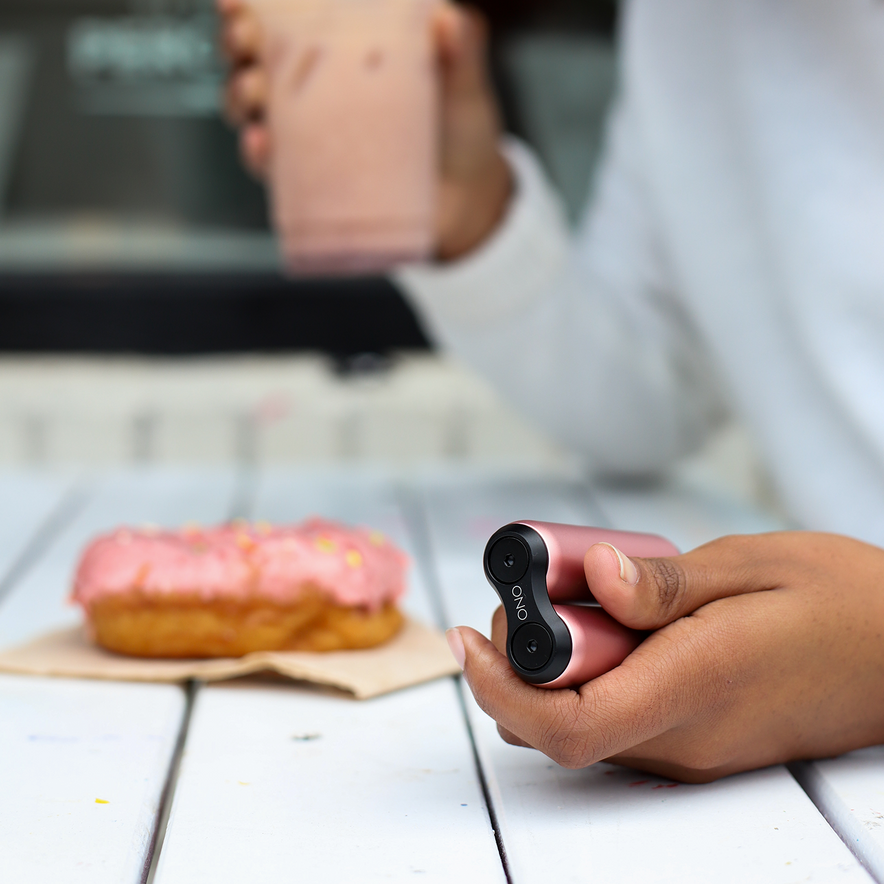 A person holds a pink ONO Roller Aluminum in one hand and a pink drink in the other, next to a pink frosted donut on a white wooden table. The sleek aluminum design makes it perfect for fidgeting while enjoying your treat.