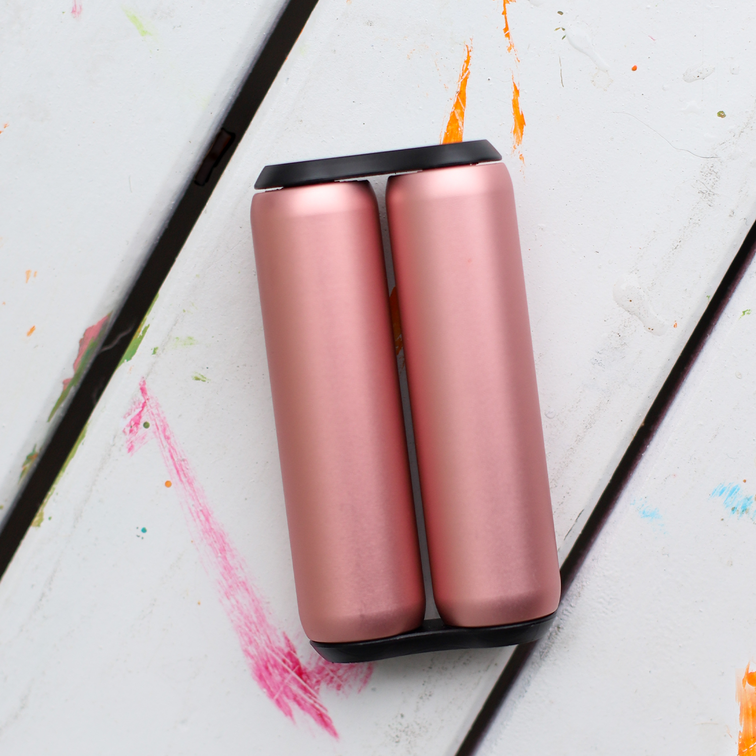 Two ONO Roller Aluminum canisters in rose gold with black lids sit side by side on a white wooden surface splattered with colorful paint.