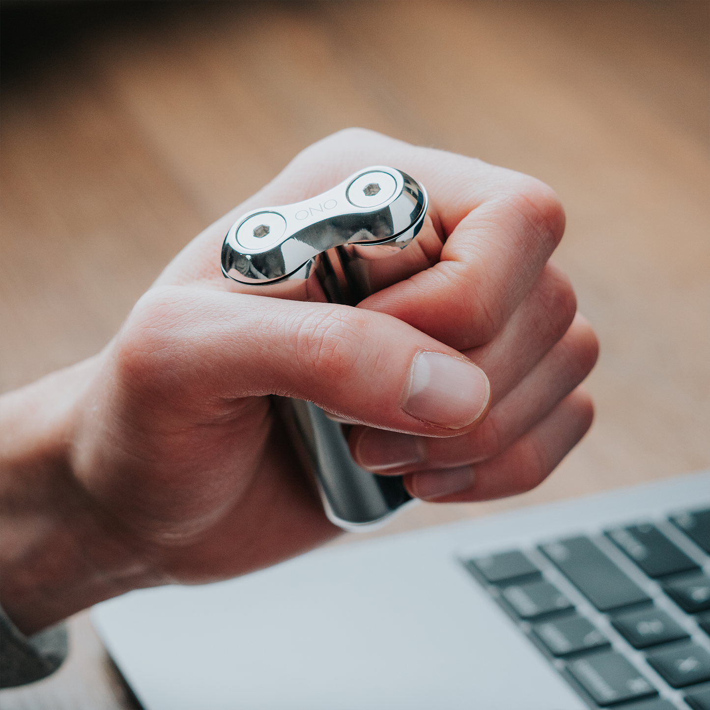 A hand holds the small, shiny ONO Roller Steel above a laptop keyboard on a wooden surface. The device features two circular parts and is engraved with ONO.