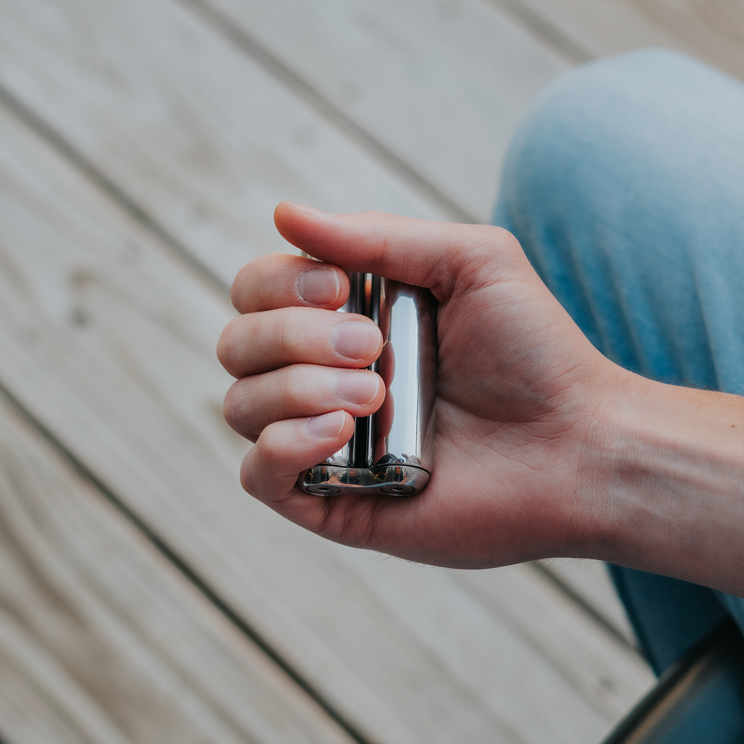 A hand grips a gleaming ONO Roller Steel fidget accessory, with a wooden deck and part of blue pants visible in the background.