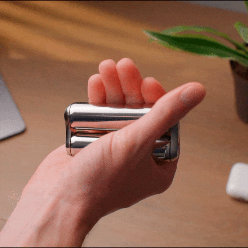 A hand holds a shiny ONO Roller Steel, split into two to reveal a hidden compartment. A plant and computer accessories blur in the background, highlighting this stylish fidget accessory as unique desk decor.