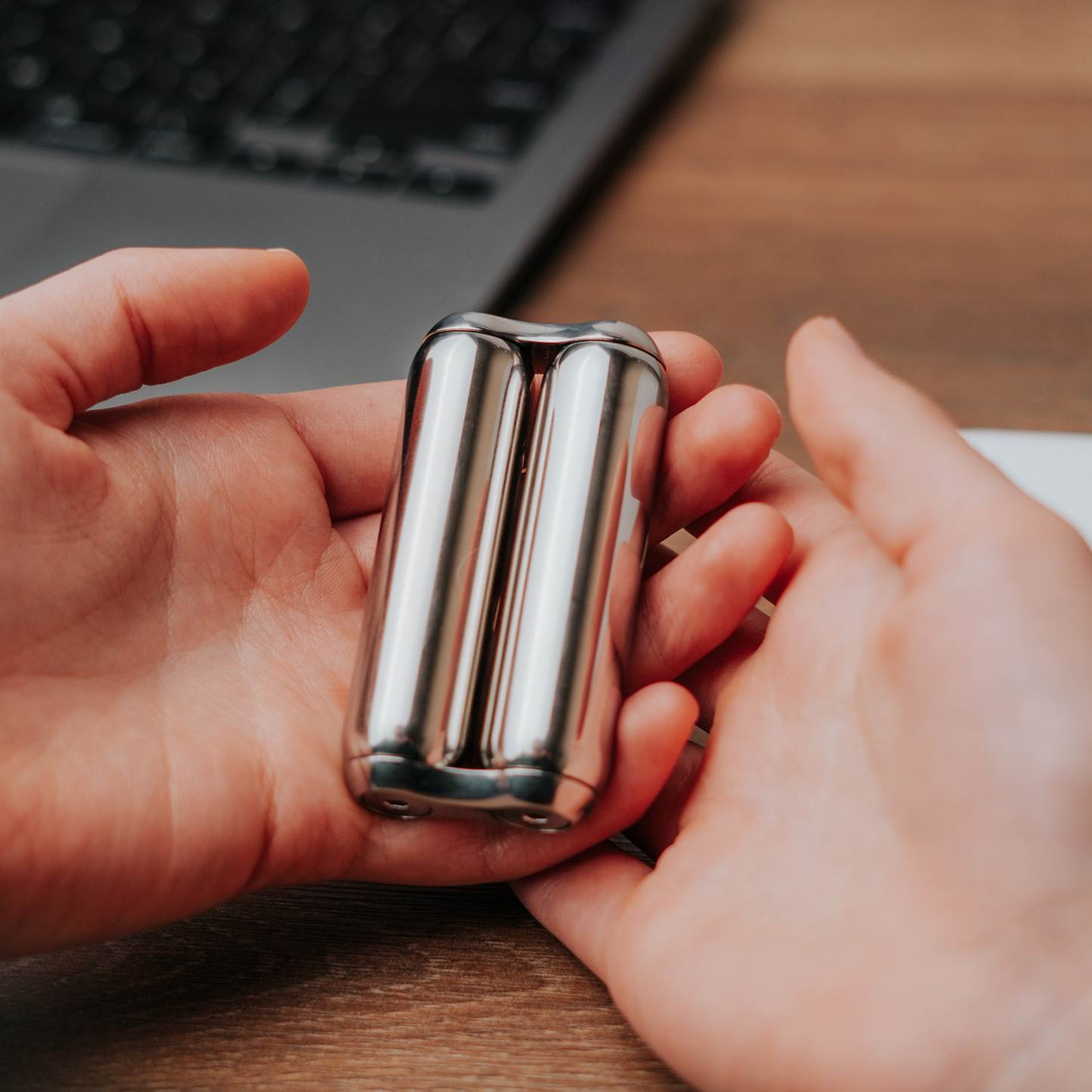 A person holds a compact, silver ONO Roller Steel hand massager in their palms, with a laptop on a wooden desk in the background—ideal as a fidget accessory or stylish desk decor.