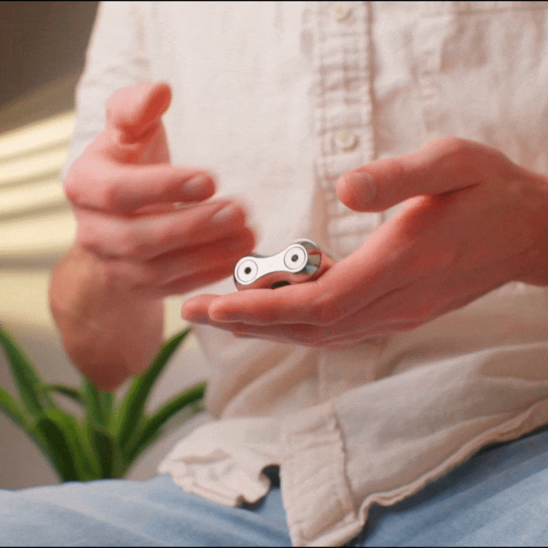 A person in a light-colored shirt spins the ONO Roller Steel fidget accessory with both hands, while a green plant appears blurred in the background.