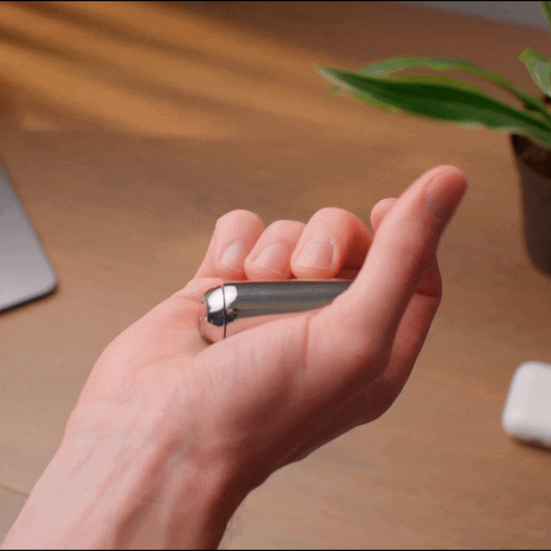 A hand flips a sleek ONO Roller Steel fidget accessory with the thumb. In the background, a blurred plant, part of a laptop, and a wireless earbud case rest on a wooden desk.