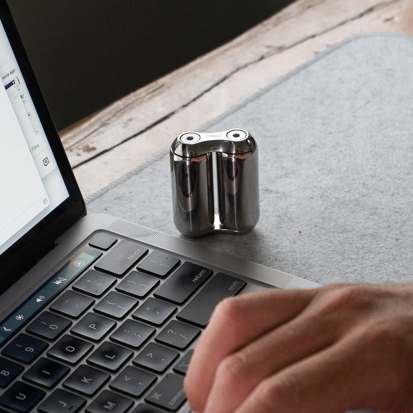 A hand uses a laptop on a gray mat next to an ONO Roller Steel—a sleek, metallic, capsule-shaped fidget accessory with two round openings—adding modern style to the wooden desk.