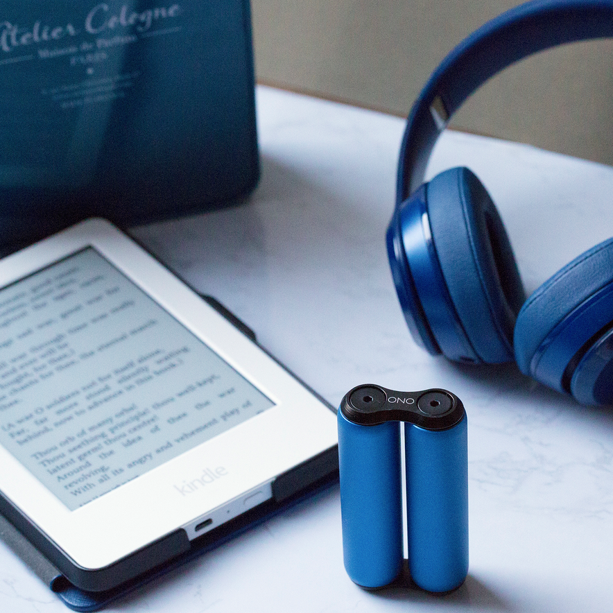 A blue Kindle e-reader, blue over-ear headphones, an ONO Roller Aluminum fidget toy, and a blue Atelier Cologne box are arranged on a white marble surface.
