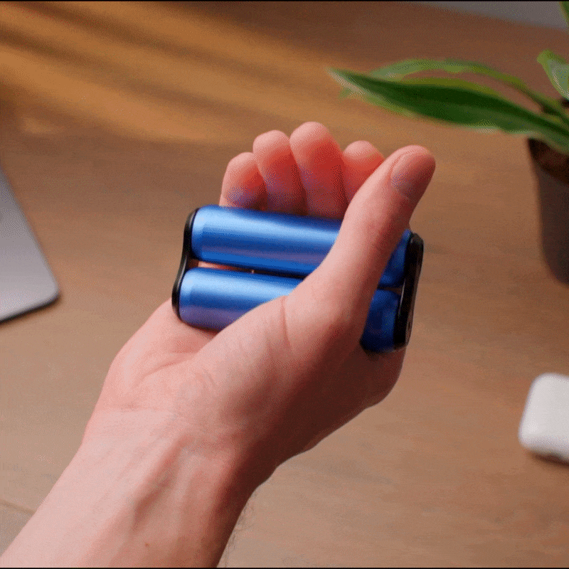 A hand squeezes and rolls the ONO Roller Aluminum fidget toy, featuring two cylindrical bars, on a wooden desk with a potted plant and electronic device nearby—ideal for fidgeting anytime.