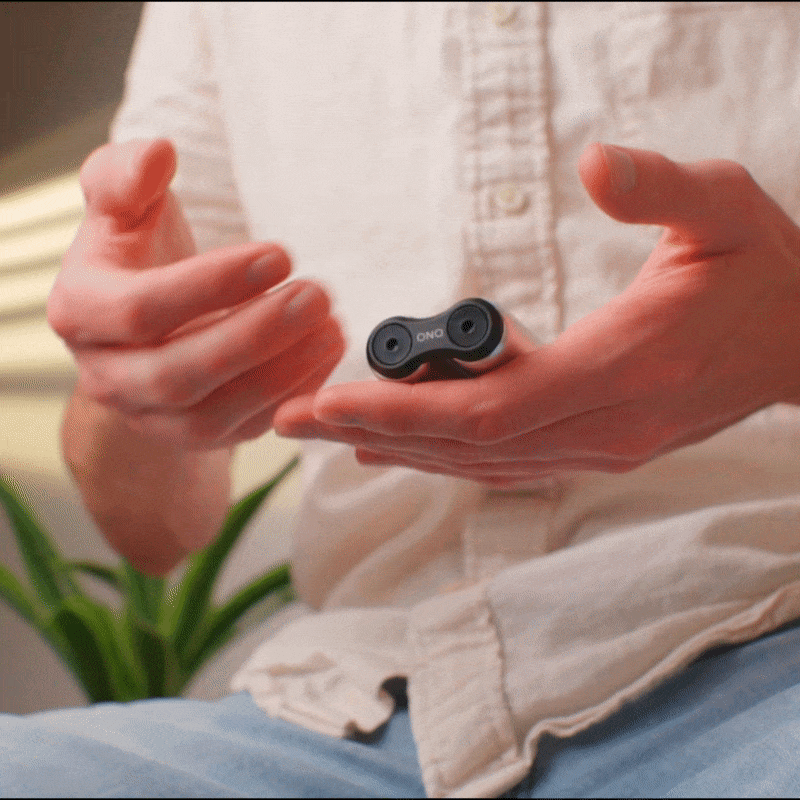 A casually dressed person fidgets indoors with the ONO Roller Aluminum, a small black double-lens gadget; a blurred green plant appears in the background.