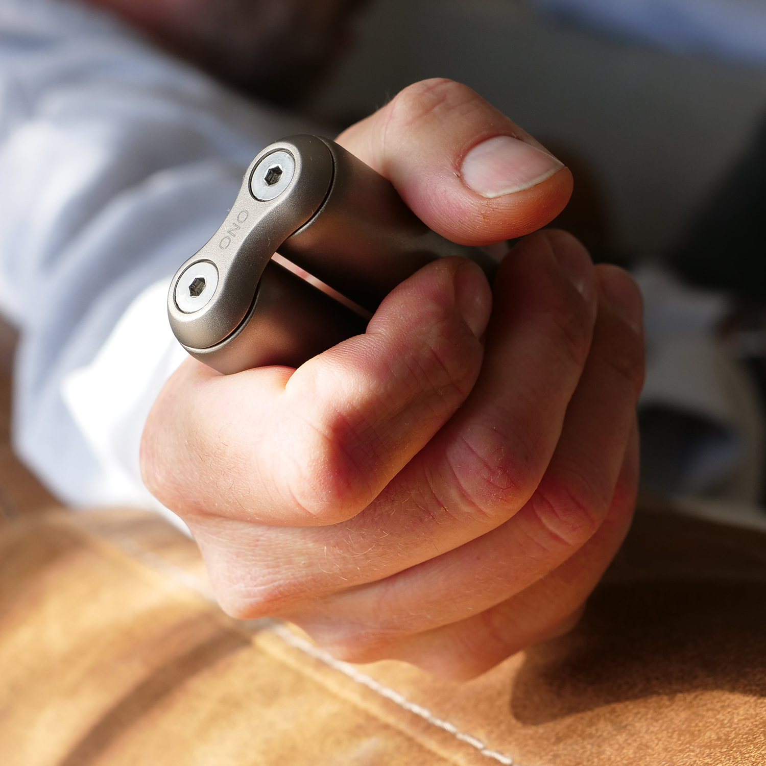 A hand holds a compact ONO Roller Titanium fidget accessory with two visible screws, shown on a tan leather surface—ideal as a unique desk decor piece.