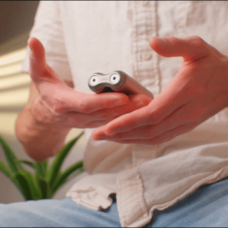 A person in a light shirt holds and demonstrates the compact, grey ONO Roller Titanium—featuring two circular buttons—possibly used as a fidget accessory. A blurred plant background enhances its appeal as stylish desk decor.