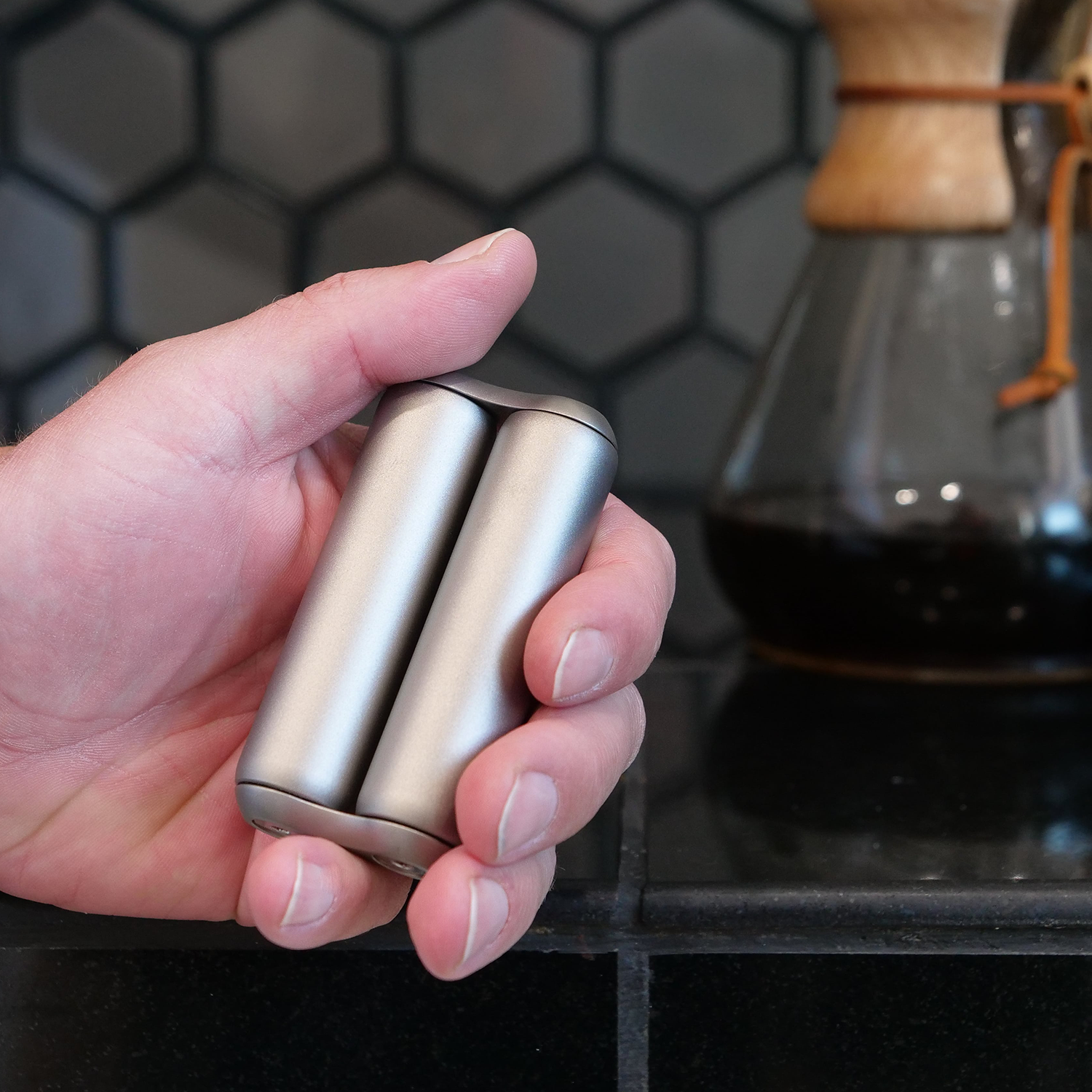 A hand holds the compact, modern ONO Roller Titanium grinder set in front of a coffee pot and hexagonal tile backsplash—ideal as stylish desk decor.
