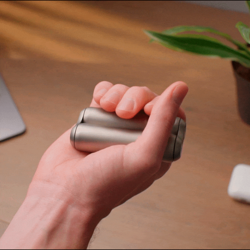 A hand smoothly rolls two ONO Roller Titanium fidget rollers between the fingers, with a blurred desk and potted plant in the background—ideal as a calming accessory or stylish desk decor.