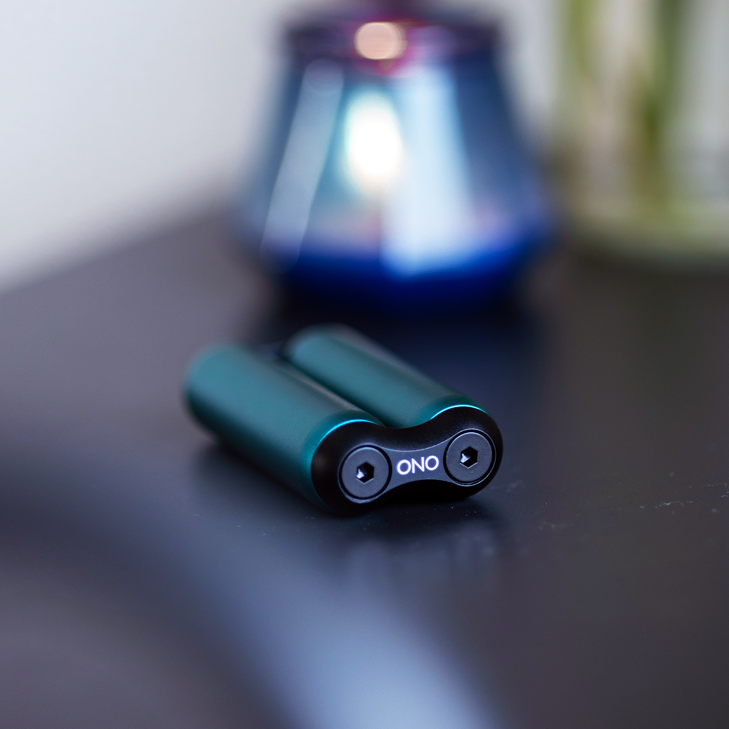 A compact black ONO Roller Aluminum with sleek aluminum accents sits on a dark surface, while a blurred blue and silver object appears in the background—ideal for discreet fidgeting.