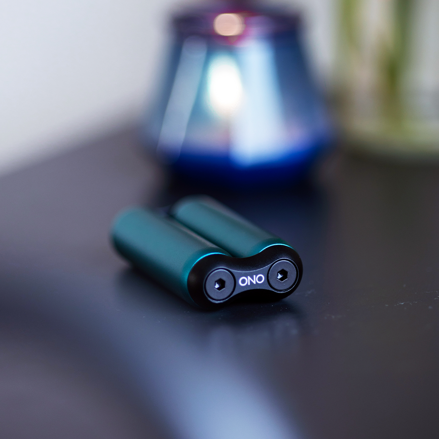 A compact black ONO Roller Aluminum with sleek aluminum accents sits on a dark surface, while a blurred blue and silver object appears in the background—ideal for discreet fidgeting.