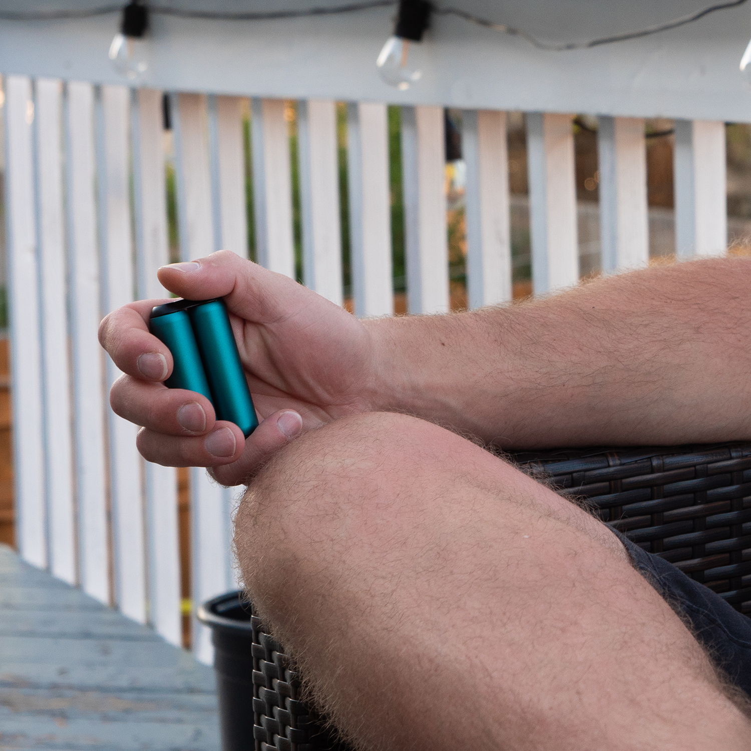 A person with light skin sits outdoors on a chair, fidgeting with an ONO Roller Aluminum in their hand. A white fence and string lights can be seen in the background.