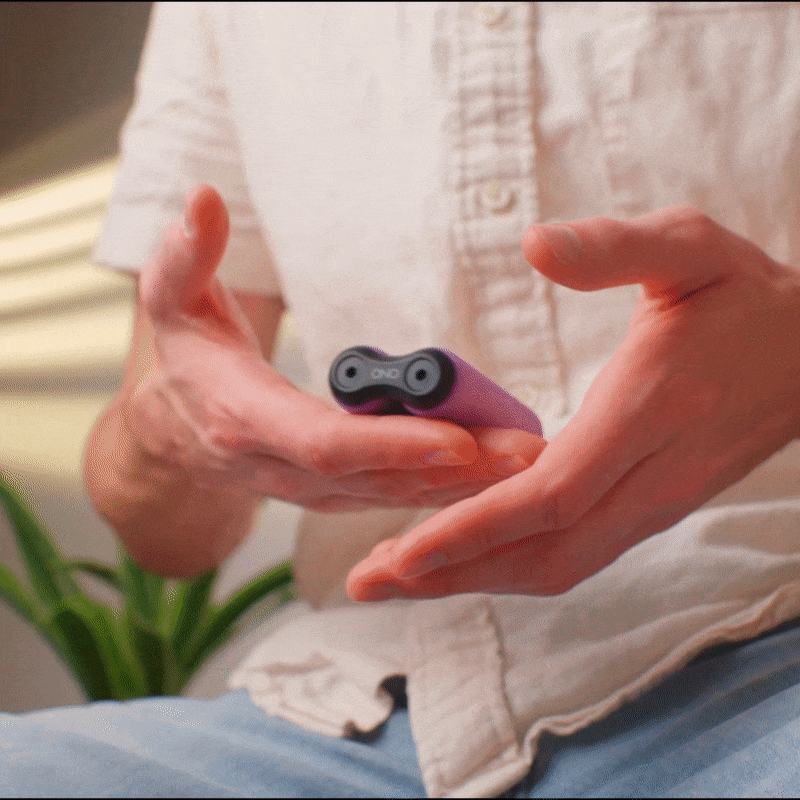 A person in a light shirt holds an ONO Roller Soft Touch Plastic with both hands indoors, a green plant behind them. The purple stress-relief device is centered and shown close up in their hands.