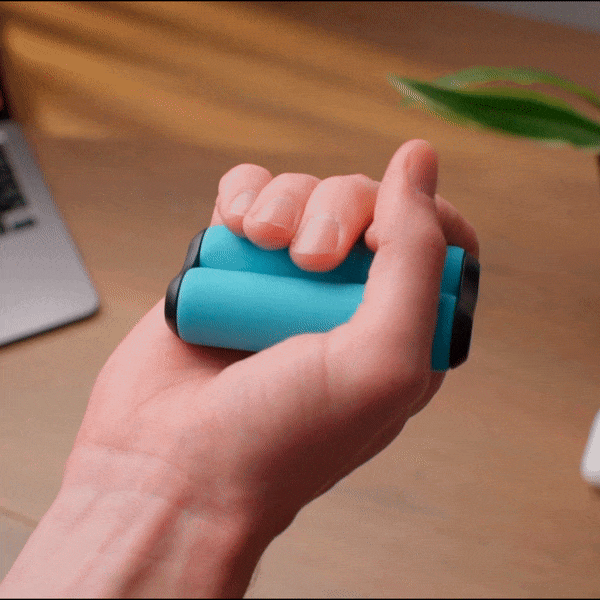 A hand squeezes the ONO Roller Soft Touch Plastic, a blue cylindrical silent fidget toy with black ends, on a wooden desk near a laptop and green plant.