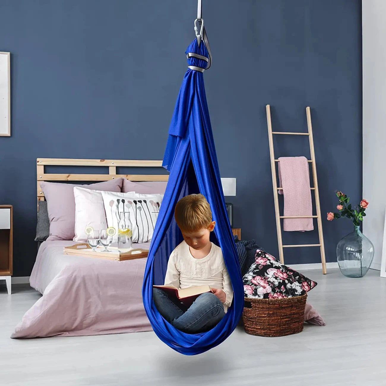 In a cozy bedroom, a child reads in an Extra-Large Sensory Swing Hammock for Kids and Adults with Special Needs. The room has pink-bedded bed, ladder shelf with pink towel, floral cushion on the floor, and dark gray walls enhancing the therapeutic ambiance.