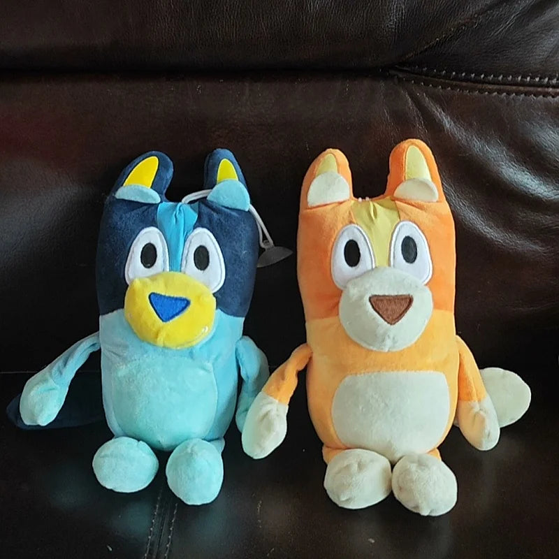Two 17cm Bluey and Bingo Family Plush Toys, soft stuffed cartoon dog dolls with large eyes and friendly expressions, sit side by side on a dark brown leather couch. Perfect for birthday or Christmas gifts.