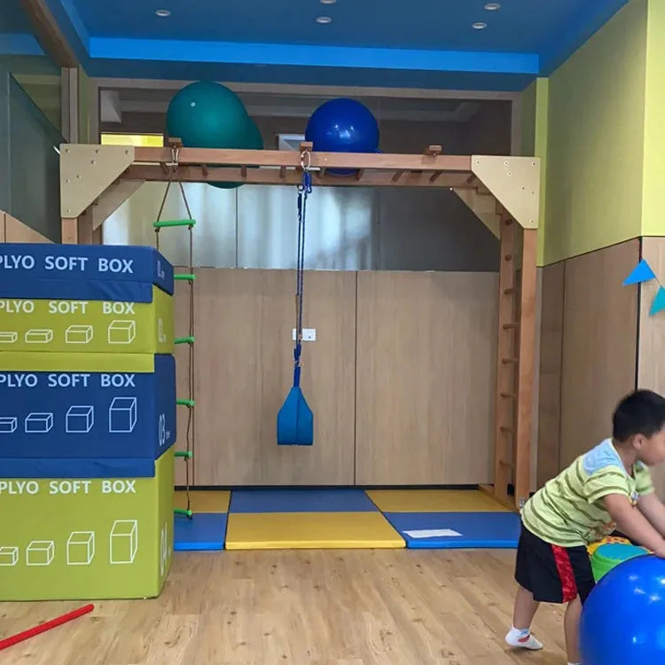In an indoor gym, a child plays with the Indoor Sensory Fitness Ladder for Kids, featuring colorful soft boxes on the left and a central wooden climbing apparatus. Blue exercise balls enhance sensory integration, while mats cover the floor for safety and fun.