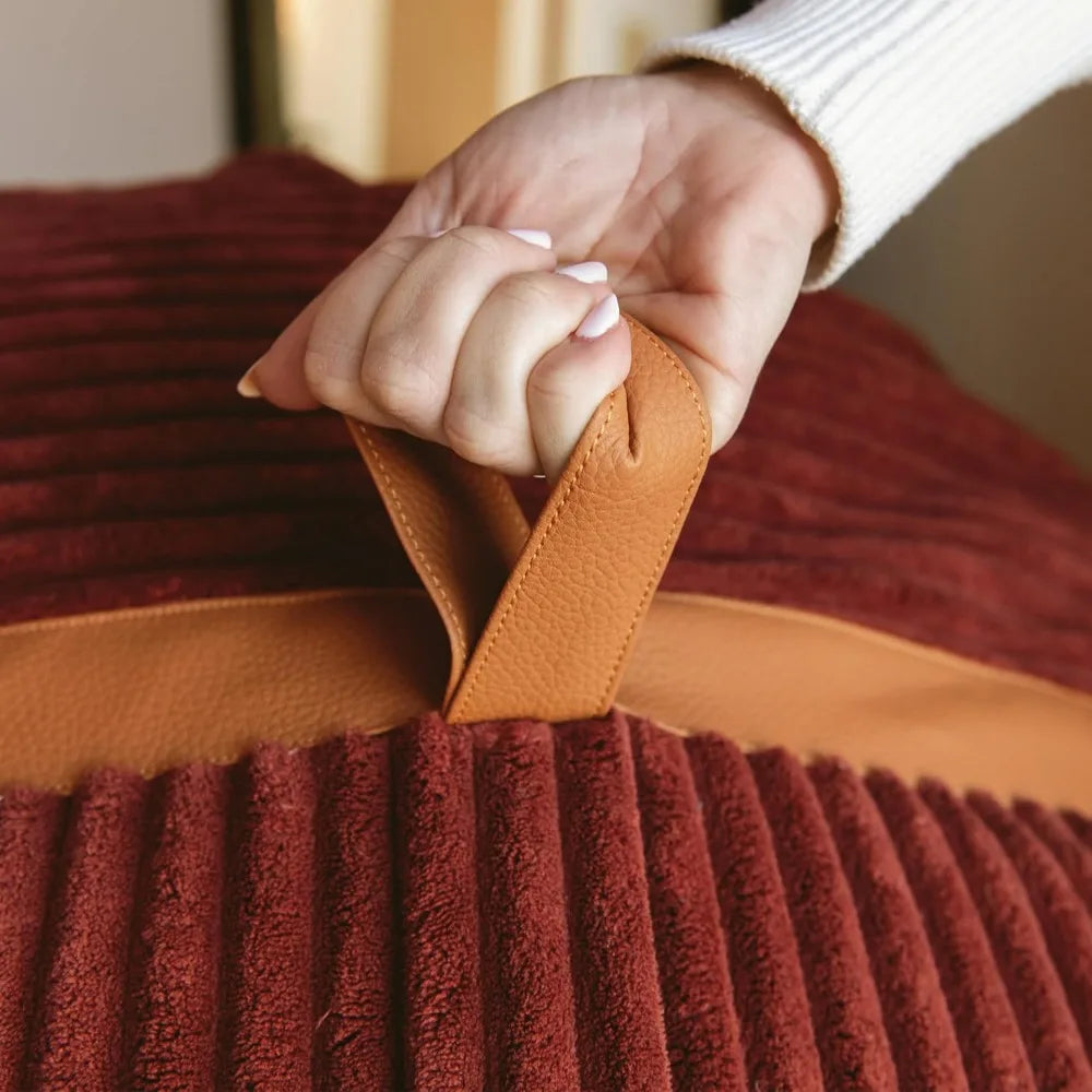 A hand with light-colored nails grips the tan leather handle of the Fuf XXL Foam Filled Bean Bag Chair in Deep Terracotta, a 6-foot plush cushion.