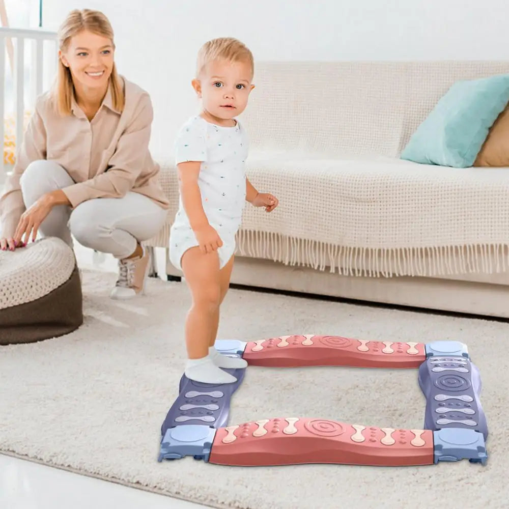 A toddler balances on the Sensory Balance Beam Training Board for Kids, a stackable toy enhancing coordination and motor skills, on a carpeted floor. Nearby, a woman kneels with a supportive smile amidst a cozy room with a sofa and cushions in soft tones.