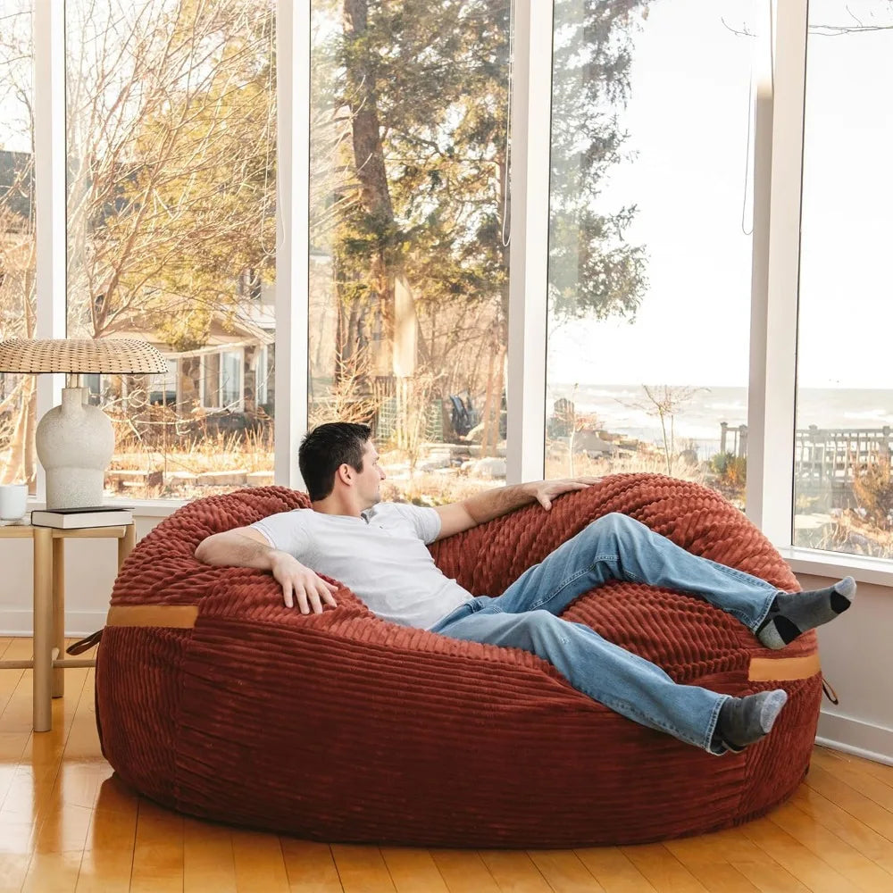 A man in a white shirt and jeans relaxes on the Fuf XXL Foam Filled Bean Bag Chair Deep Terracotta 6 Feet in a bright room with large windows, gazing outside at trees and a water view.