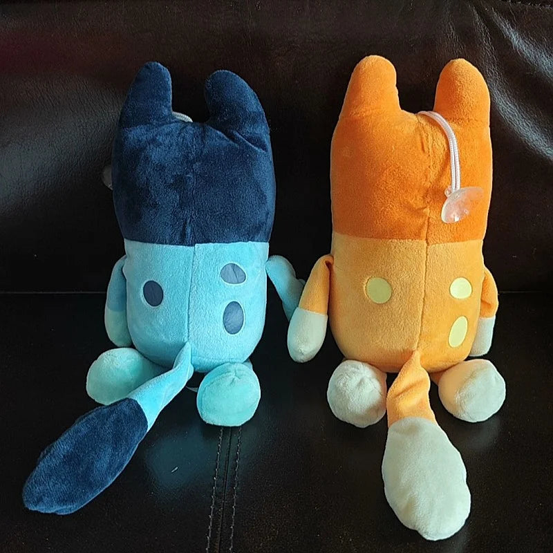 Two Bluey and Bingo Family Plush Toys, one blue and one orange, sit side by side on a dark surface with their backs to the camera. The orange plush, perfect for gifts, features a pacifier attached to its head. Sizes: 17cm/28cm/40cm.