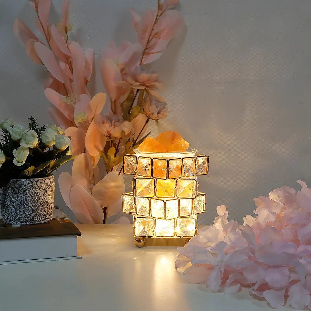 The Himalayan Salt Crystal LED Lamp - USB Night Light for Home Décor and Relaxation radiates a warm ambiance on a table with pink and white flowers, a patterned pot of small white blooms, and a book, creating a cozy space with its convenient USB plug.