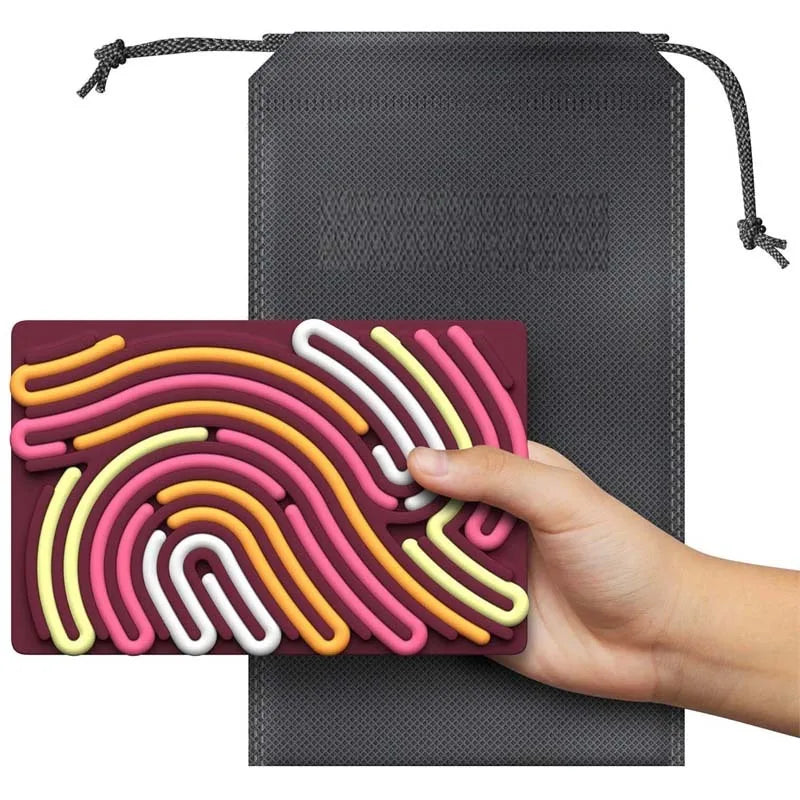 A hand holds the red Childrens Puzzle Silicone Sensory Activity Board—food grade, BPA free—with colorful raised squiggly lines; a black drawstring pouch in the background makes it a perfect decompression tool for sensory play.