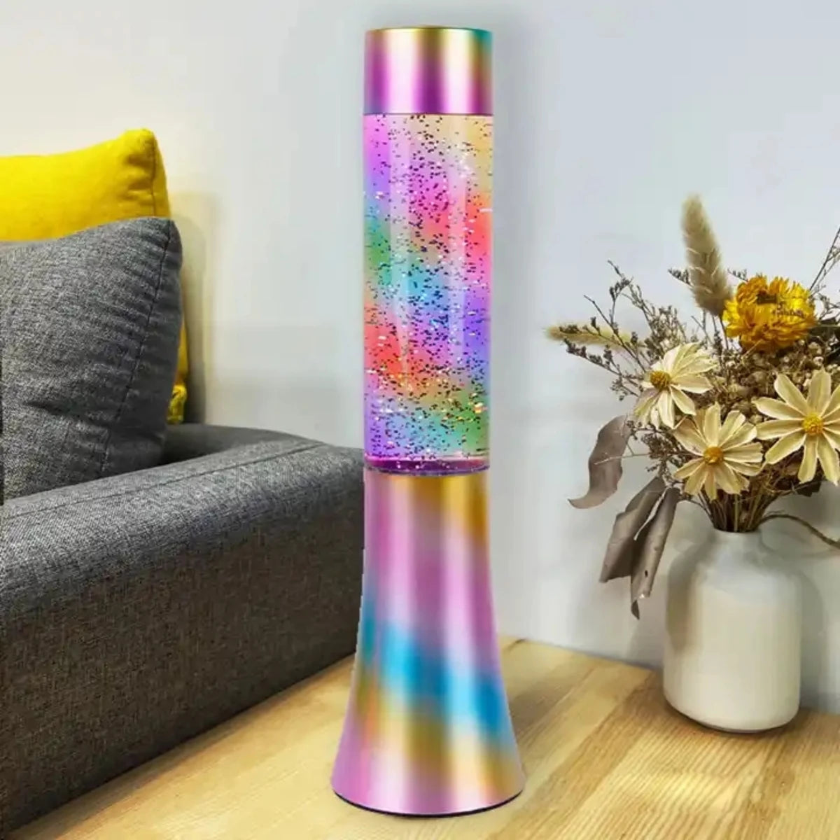 A 13-inch Color-Changing LED Glitter Lava Lamp brightens up a wooden floor by a gray sofa with a yellow cushion, adding vibrancy to the decor. Nearby, a white vase with dried flowers rests against the light wall.