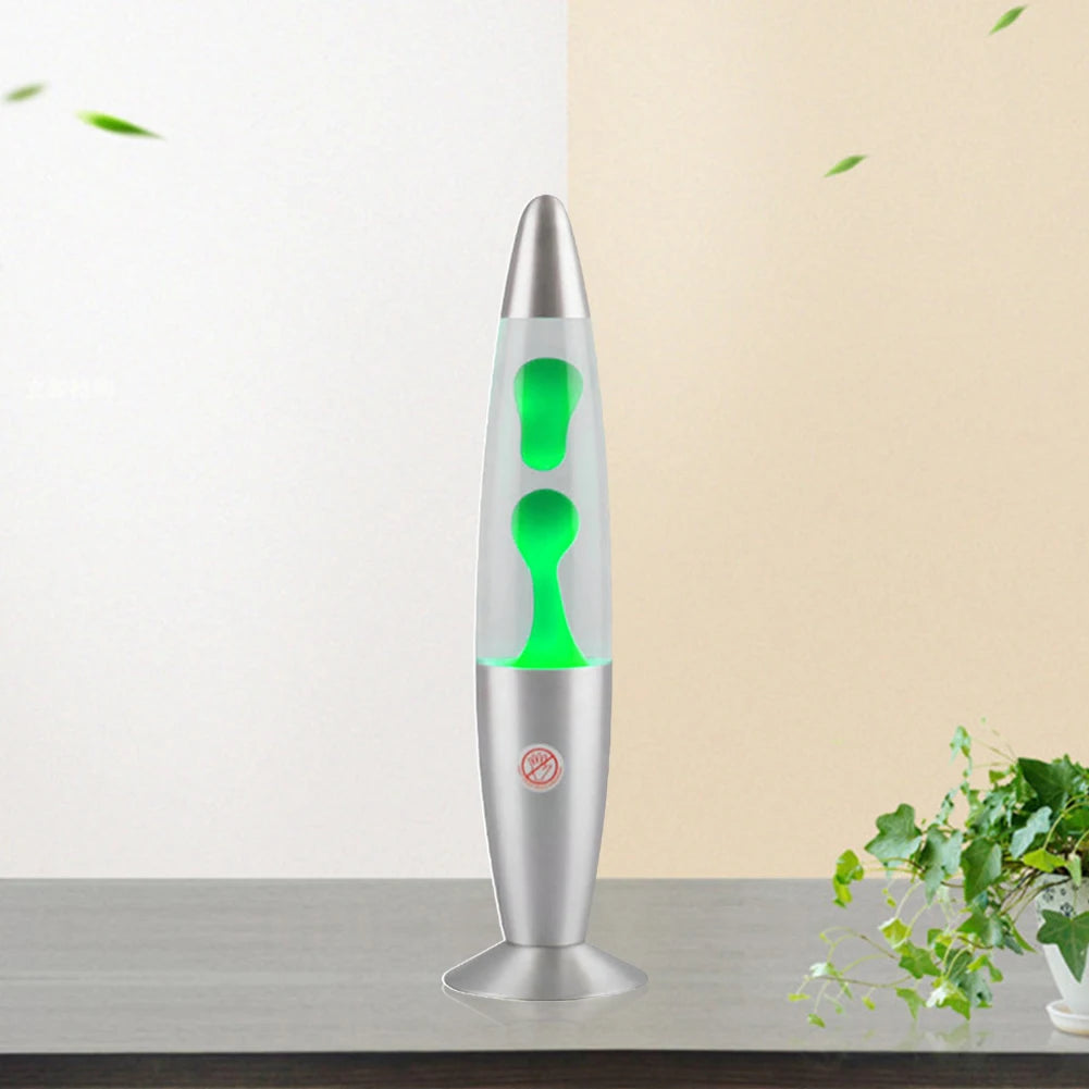 A Luminous Volcano Lava Jellyfish LED Night Light casts a gentle glow with bright green wax on a gray surface. It stands against a two-toned wall, beige to the left and light green to the right, and a small plant with lush leaves graces the bottom right corner.