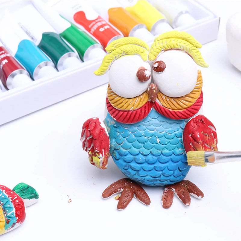 A colorful clay owl figurine crafted from the No-bake Air-dried Shaping DIY Plasticine Clay Set with 12 Colors of Paint features a blue body, red wings, and yellow head feathers. In the background, a variety of paint tubes await and a second smaller painted owl is nearby.