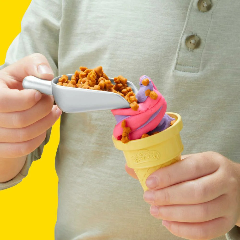 Someone is creatively using the Hasbro Play Doh Rainbow Swirl Ice Cream Playset, forming a toy ice cream cone with pink and purple Play-Doh, and adding brown pieces with a plastic scoop against a bright yellow backdrop.