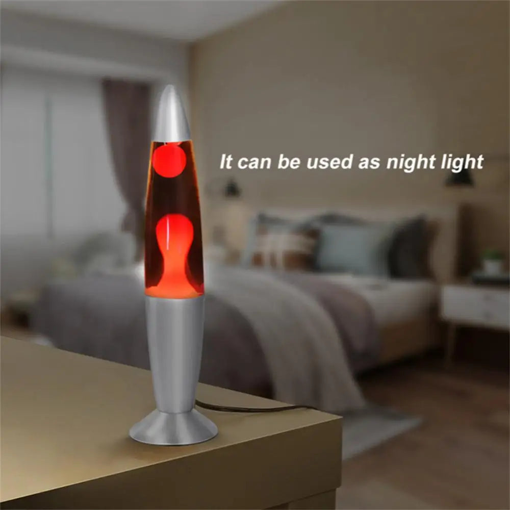 A Mesmerizing Jellyfish Lava LED Night Light, featuring a gray aluminum alloy design and red wax, adorns a bedside table in a dim bedroom. A bed with pillows and blankets forms the backdrop, suggesting its use as an ambient light for kids rooms.