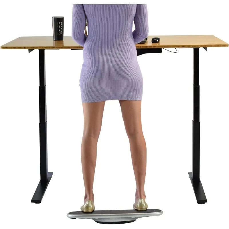 A person in a lilac dress uses the BASE Desk Standing Board – Wobble Board with Anti-Fatigue Mat at a height-adjustable desk, with a mug and mouse on top—an inspiring setup for better posture, viewed from behind.