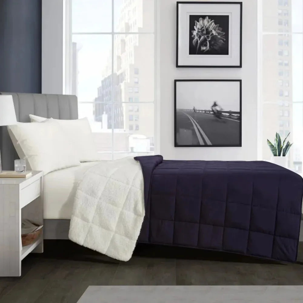 A modern bedroom features a neatly made bed with a Luxurious Microfiber Weighted Blanket in navy blue and white. Large windows offer an urban view, while monochrome art decorates the wall and a small plant sits on the bedside table.