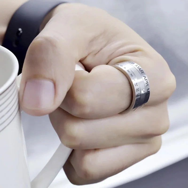 A hand holds a white mug while wearing the Stainless Steel Anxiety Ring—a silver unisex rotating calendar date fidget spinner ring engraved with months and numbers. A black wristband is also worn.