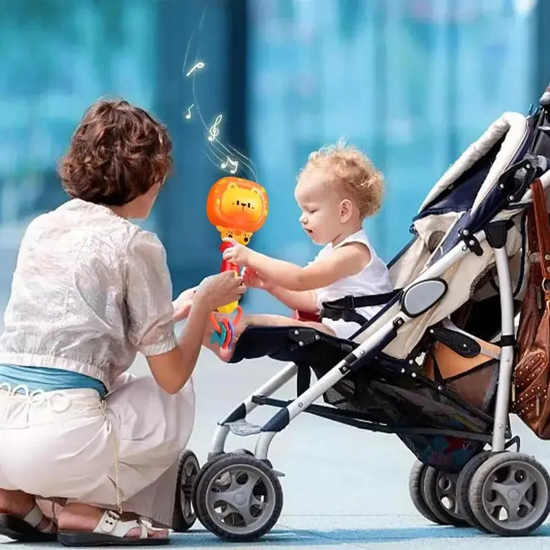 A woman crouches beside a child in a stroller who holds the Musical Teething Rattle with LED Lights – Gentle Soft Teether & Interactive Learning Toy, enjoying its glowing orange hue and soft music in an outdoor setting with a blurred background.