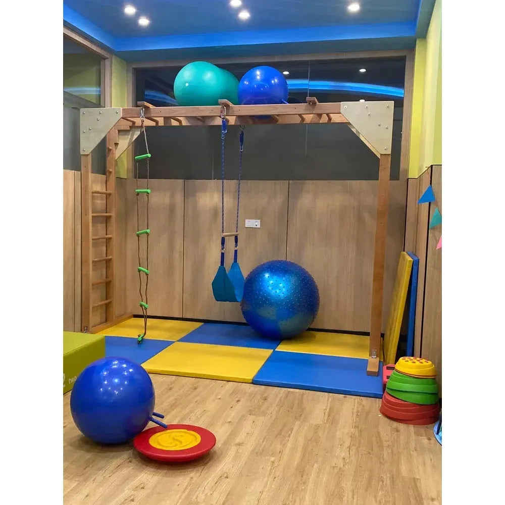 The indoor play area includes an Indoor Sensory Fitness Ladder for Kids: Swing Climbing and Suspension Play Equipment, ideal for sensory integration training. It features a wooden climbing frame, colorful gym mats, blue exercise balls, bright lighting, a wooden floor, swings, and stackable cones.