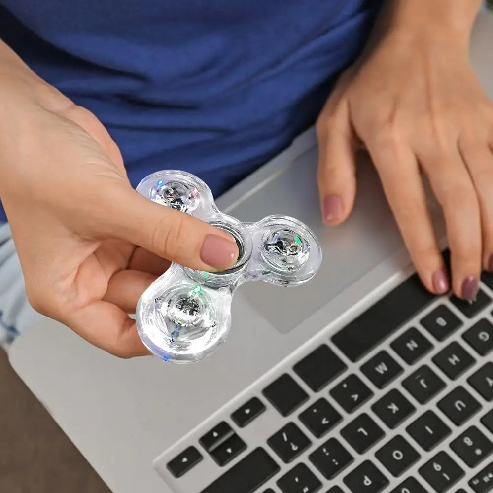 A casually dressed person holds the Crystal Finger Spinners Luminous LED Light Fidget Spinner above their laptop keyboard, capturing a stress relief moment amid a busy day.