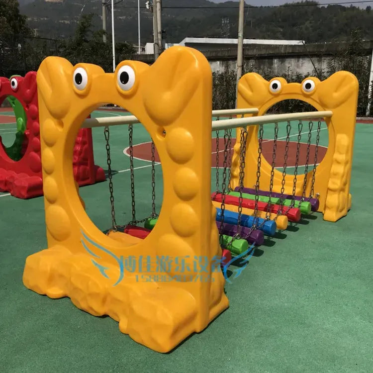 A vibrant playground set includes a yellow, frog-shaped climber with a circular cutout and multicolored planks. Complemented by metal toy elements and a suspended swinging bridge, it sits on a green cushioned surface. Suitable for ages 3-8, its perfect for unisex fun.