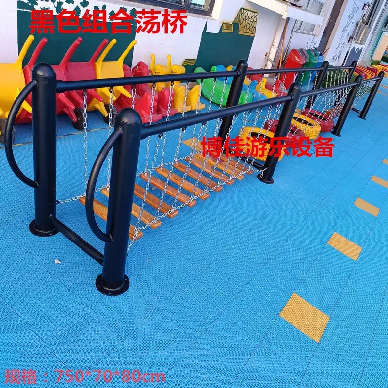 Outdoor toys including black metal-framed swinging bridges with wooden planks and chain connections sit on a blue rubber mat. Colorful seating enhances the space, offering an engaging area that complements other unisex childrens toys alongside swings and climbing nets for sensory integration.