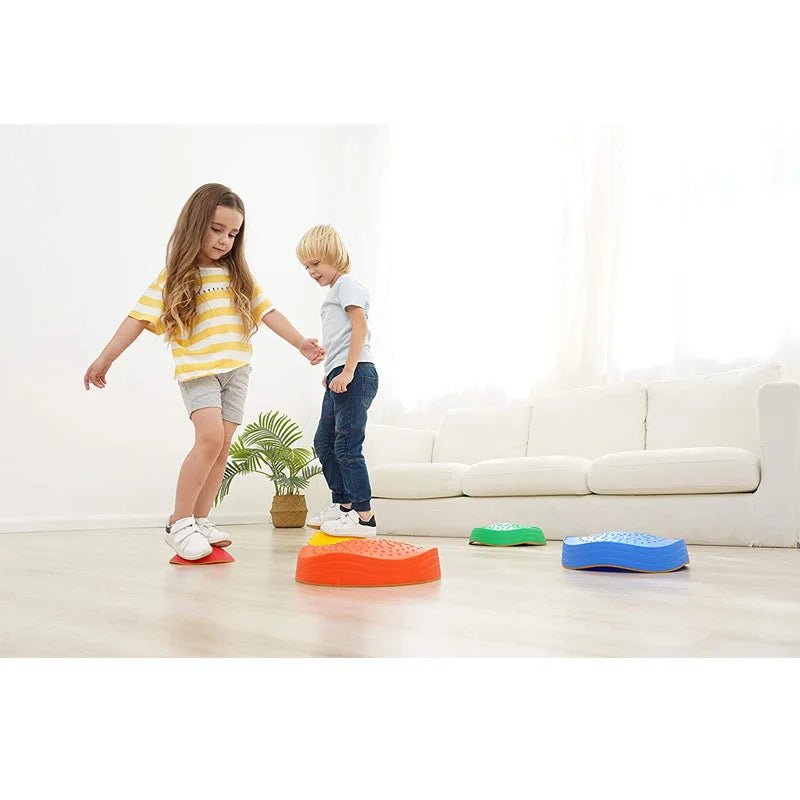 In a bright room, two children enjoy the Kids 5-Piece Outdoor Balance Stepping Stones Set for Coordination and Obstacle Play on a wooden floor. The girl in a yellow striped shirt balances on a red stone, weaving through the course, as the boy in a white shirt observes. A white sofa and plant are visible in the background.