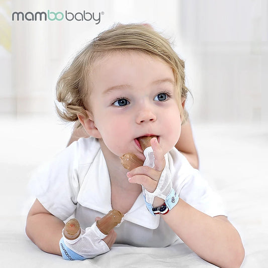 A baby with light brown hair, in a white shirt, is on a bed wearing Mambobaby Anti-Nail Biting Gloves with silicone cots to curb thumb sucking. The mittens feature brown details, set against a softly focused indoor background.