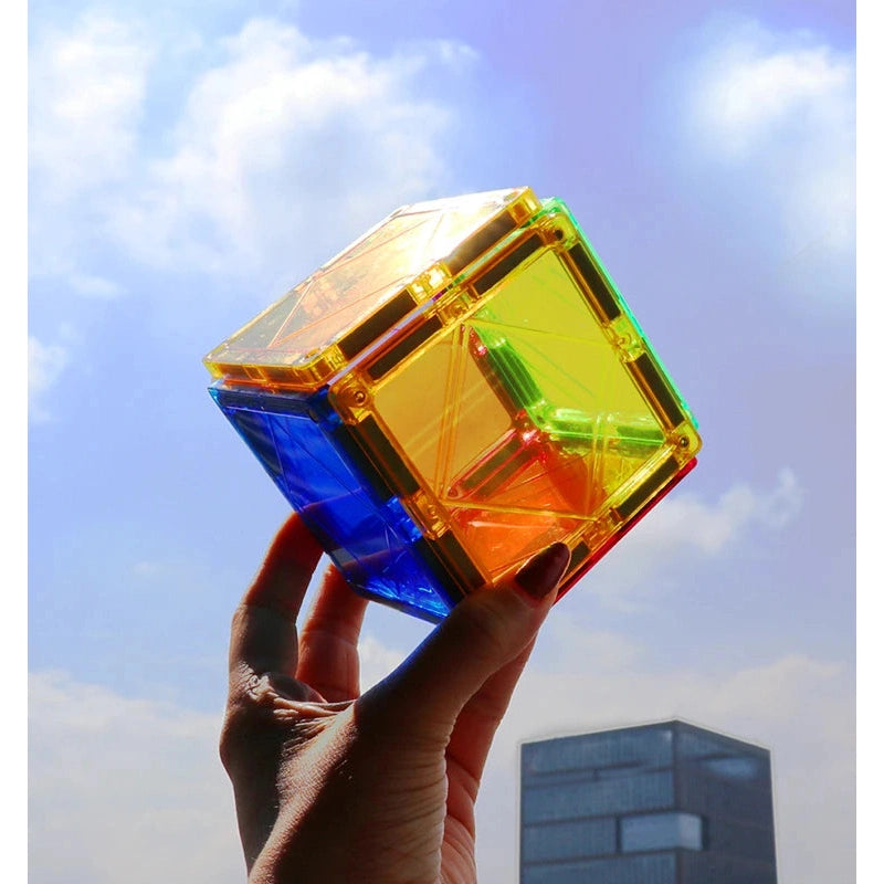 A hand holds a transparent, colorful cube made of Magplayer Magnetic Construction Blocks against a bright sky. Ideal for STEM learning, its yellow, orange, red, and blue panels sparkle creatively as a building stands in the background.